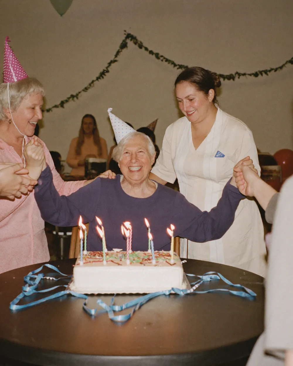 An older woman beaming at her birthday celebration as friends clap around a cake