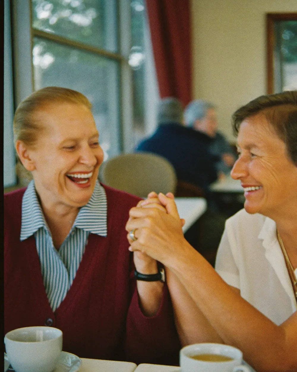 Two older women laughing and holding hands over tea